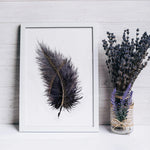 L'image montre un cadre blanc contenant une illustration de plume noire avec des points dorés, ainsi qu'un bouquet de fleurs de lavande dans un vase en verre sur une surface en bois.