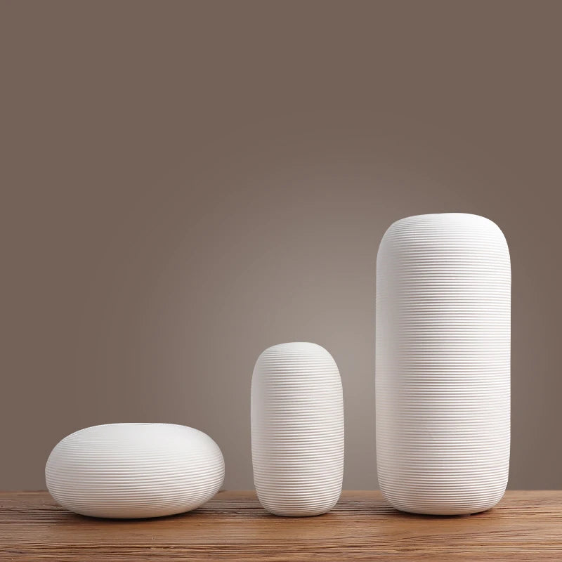 Vases blancs cylindriques de différentes tailles, posés sur une table en bois, contre un fond neutre marron.