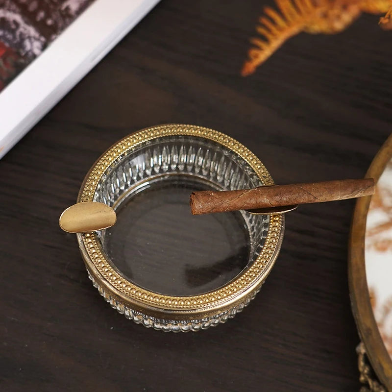 Un cendrier de style vintage en verre et métal doré, avec un cigare posé sur le côté.