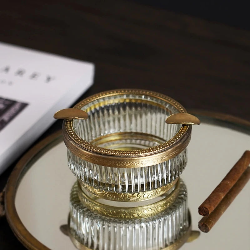 Un élégant cendrier en verre et métal doré, avec des détails ornés, posé sur une surface sombre.