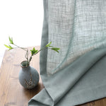 Voilage en tulle gris transparent tombant délicatement sur une table en bois avec un petit vase bleu et des branches décoratives.