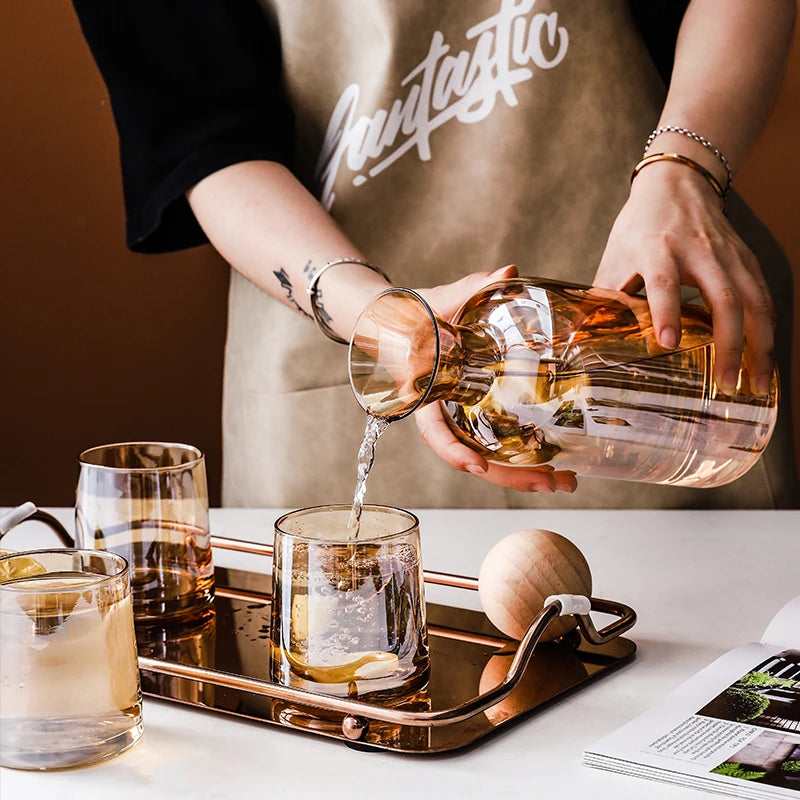 Une bouteille de verre contenant un liquide ambre est versée dans des verres sur un plateau métallique, accompagnée de tranches de fruits et de documents.