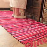 Tapis artisanal multicolore rayé rouge et rose, posé au sol dans une pièce avec un meuble en bois et une personne debout sur le tapis portant des chaussures beiges.