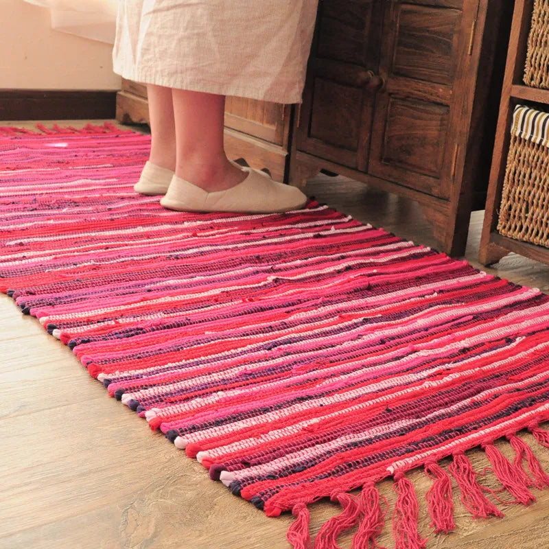 Tapis artisanal multicolore rayé rouge et rose, posé au sol dans une pièce avec un meuble en bois et une personne debout sur le tapis portant des chaussures beiges.