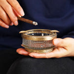 Un cendrier vintage orné de motifs dorés, contenant un cigare allumé dans les mains d'une personne.