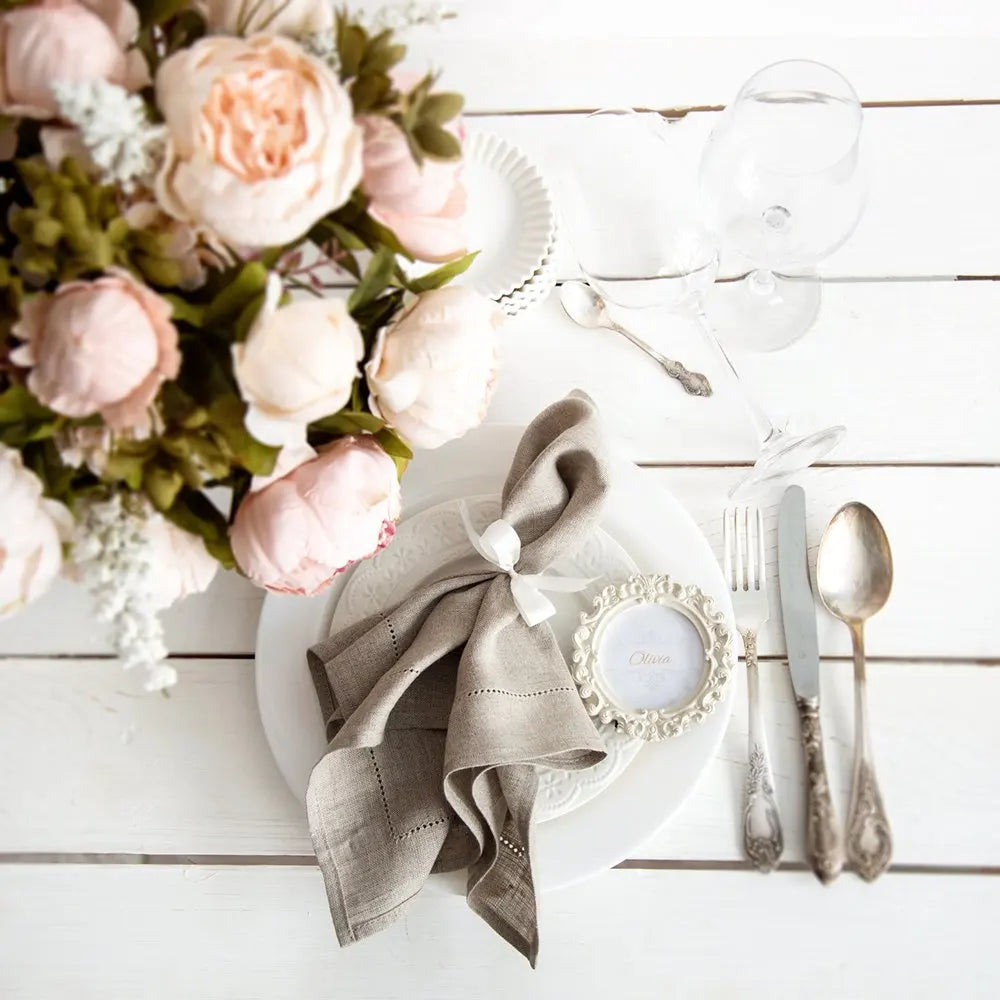 Une nature morte élégante avec des fleurs de pivoine rose pâle, des couverts en métal et une serviette nouée dans un style rustique sur un fond en bois blanc.