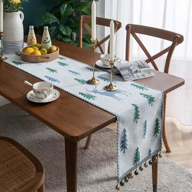 Une table en bois avec une nappe décorative au motif de forêt et de rennes, accompagnée d'accessoires de décoration, d'un bol de fruits et d'une tasse et soucoupe.