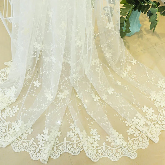 Détail du voilage blanc brodé de fleurs, étendu au sol avec ourlet festonné élégant, capturant la lumière naturelle sur carrelage clair.