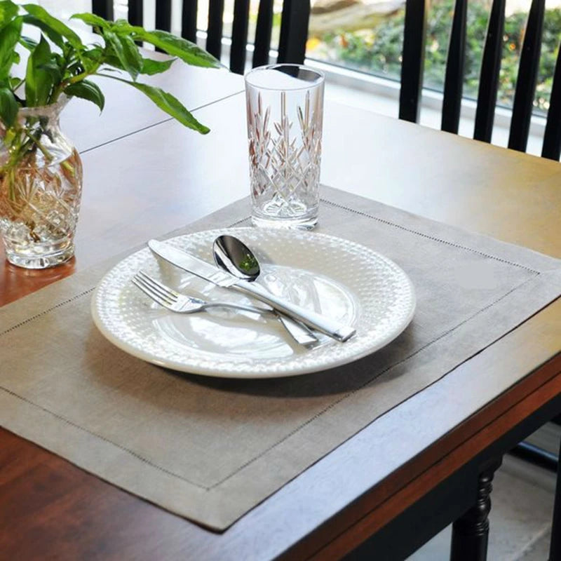 Une table dressée avec une assiette, des couverts et un verre de cristal décoré sur un chemin de table gris, au milieu d'un décor de verdure.