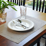 Une table dressée avec une assiette, des couverts et un verre de cristal décoré sur un chemin de table gris, au milieu d'un décor de verdure.
