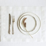 Sur une nappe blanche se trouvent une assiette et des couverts, ainsi qu'un élément décoratif naturel évoquant les plumes.