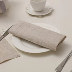 Une serviette de table en lin naturel légèrement froissée, posée sur une assiette blanche sur une surface blanche avec une tasse et une soucoupe en porcelaine.