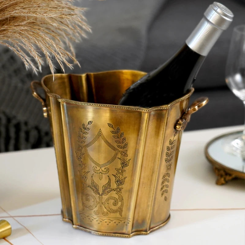 Un seau à vin doré orné de motifs floraux, contenant une bouteille de vin, sur une table blanche avec des épis de blé.