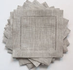 Serviettes de table en lin gris clair avec bordure piquée délicate, empilées sur un fond blanc, créant une texture et une atmosphère élégantes.