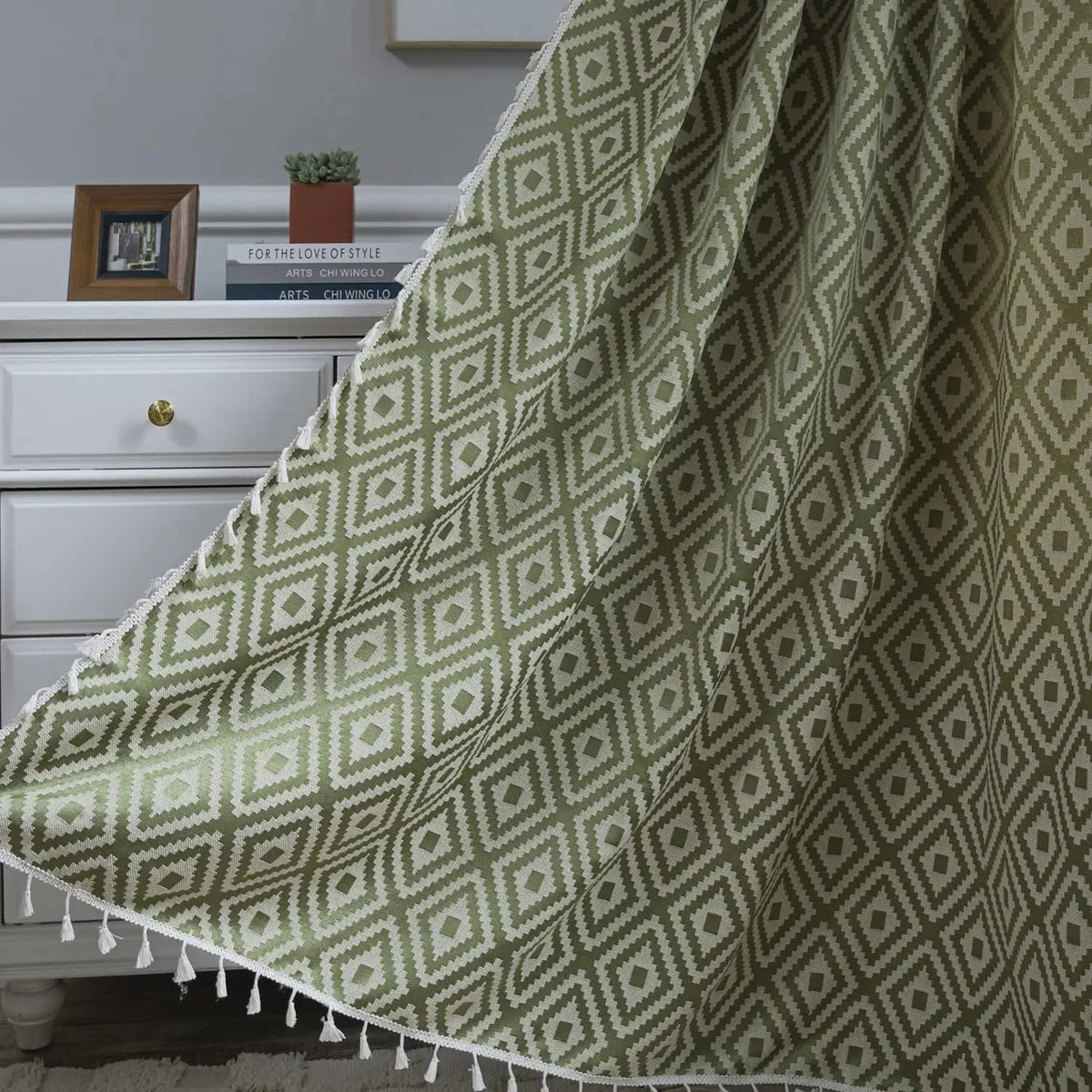 Rideau vert olive tamisant avec motifs géométriques losanges et pompons décoratifs, suspendu devant une commode blanche dans un intérieur chaleureux.