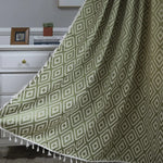 Rideau vert olive tamisant avec motifs géométriques losanges et pompons décoratifs, suspendu devant une commode blanche dans un intérieur chaleureux.