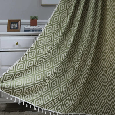 Rideau vert olive tamisant avec motifs géométriques losanges et pompons décoratifs, suspendu devant une commode blanche dans un intérieur chaleureux.
