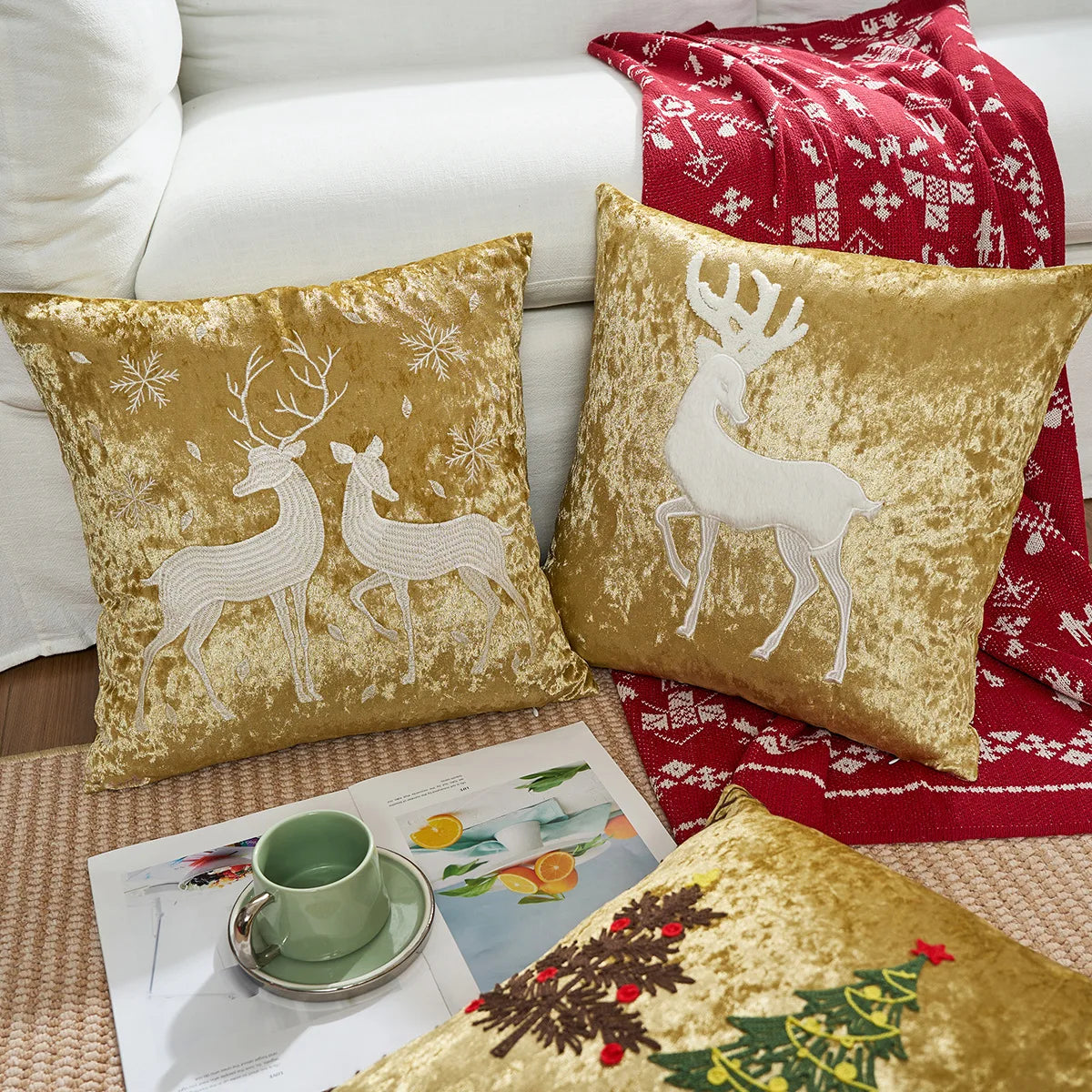Coussins dorés avec motifs hivernaux de cerfs et de flocons de neige, accompagnés d'un plaid en laine rouge à motifs nordiques, le tout disposé sur une chaise blanche aux côtés d'une tasse de thé vert et d'une revue.