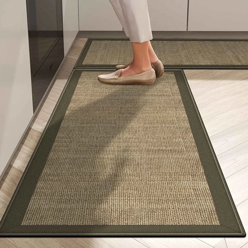 Un tapis en fibres naturelles avec une bordure de couleur sombre, posé sur un sol en bois clair, sur lequel on peut voir des pieds chaussés de mocassins beiges.