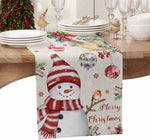 Un décor de table de Noël avec des plats, des verres et un chemin de table orné de motifs de Noël, notamment un bonhomme de neige et des boules de Noël.