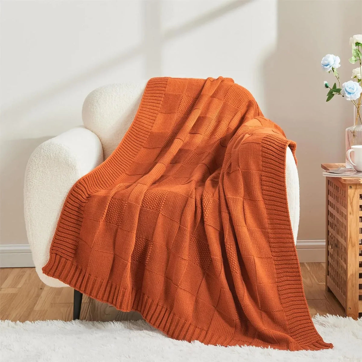 Couverture tricotée chaleureuse et luxueuse de couleur orange, posée sur un fauteuil douillet, créant une atmosphère confortable et cosy dans une pièce aux tons clairs.