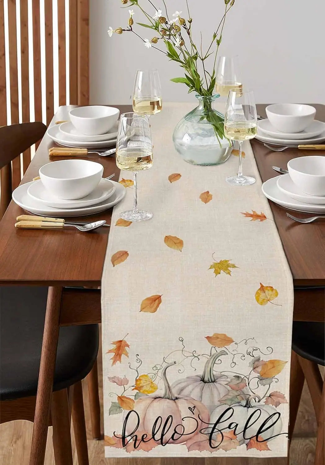 Une table de salle à manger décorée avec des verres à vin, des assiettes et une belle décoration automnale, comprenant des feuilles peintes et une citrouille ornée du texte "Hello Fall".