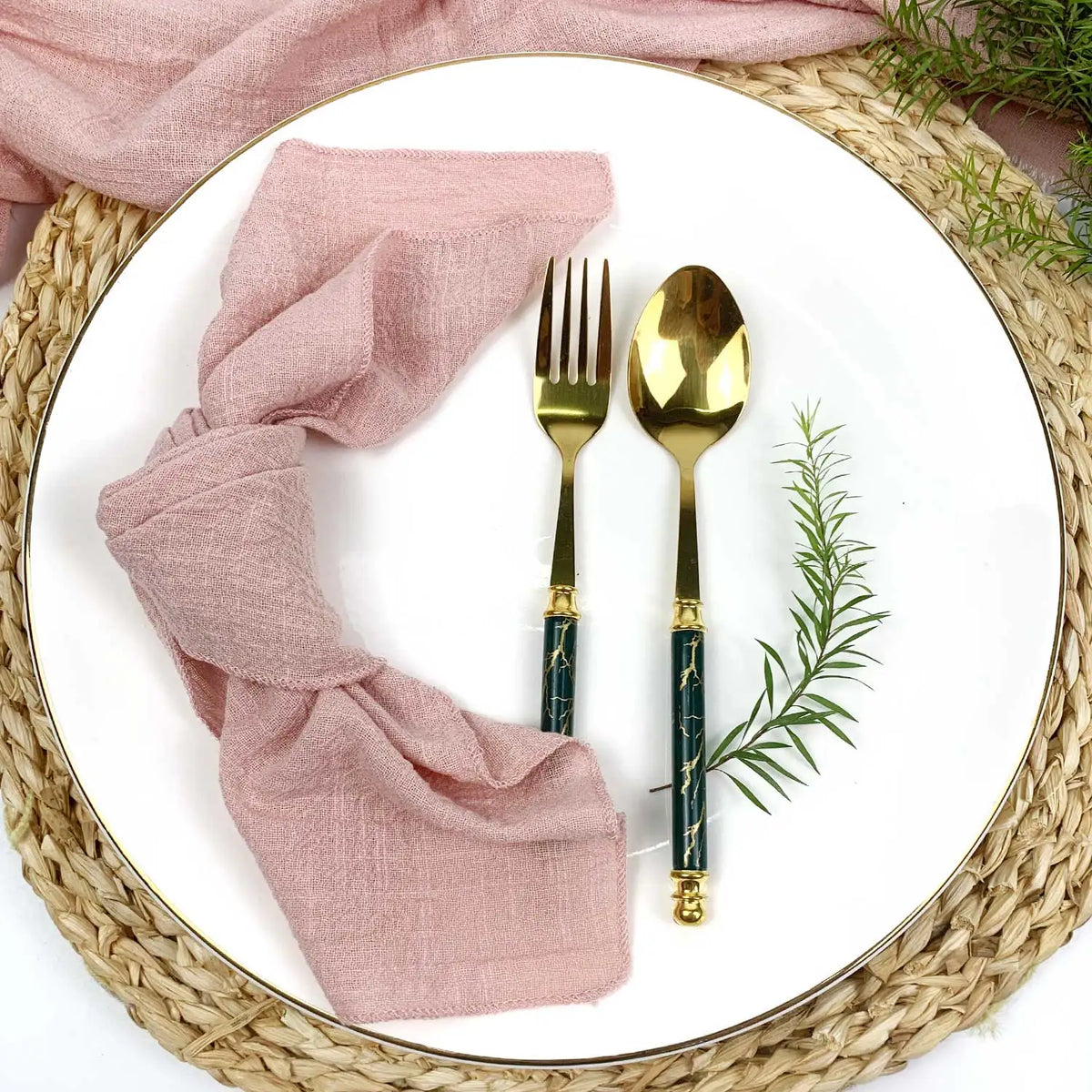 Une serviette de table rose, une fourchette et une cuillère dorées posées sur un plateau en osier, entourées de verdure fraîche.