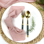 Une belle table de repas avec des serviettes roses en lin, des couverts en or et une verdure fraîche qui apportent une touche de naturel.