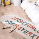 Cette image montre un tapis décoratif avec des motifs géométriques colorés et le texte "BOUGH MINSTER" imprimé au centre. Le tapis est posé sur un sol en bois clair, avec des objets décoratifs dans le coin.