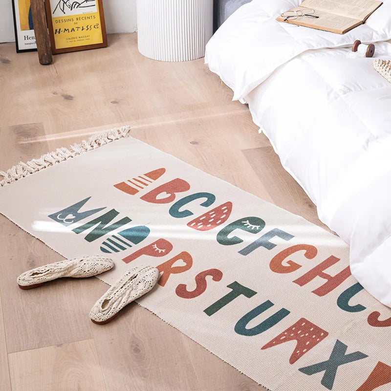 Cette image montre un tapis décoratif avec des motifs géométriques colorés et le texte "BOUGH MINSTER" imprimé au centre. Le tapis est posé sur un sol en bois clair, avec des objets décoratifs dans le coin.