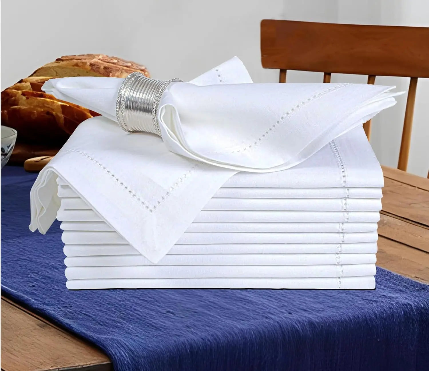 Une pile de serviettes de table blanches, pliées avec soin, déposée sur un textile bleu foncé, entourée de bols et de pains.