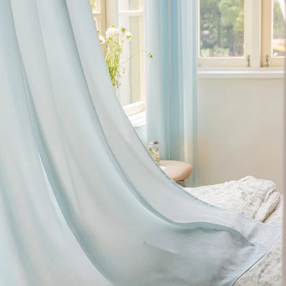 Voilage bleu translucide flottant délicatement devant une fenêtre ouverte, apportant une lumière douce à une chambre lumineuse.