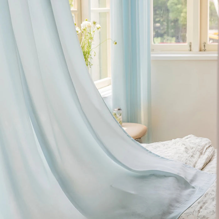 Voilage bleu translucide flottant délicatement devant une fenêtre ouverte, apportant une lumière douce à une chambre lumineuse.