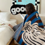 Un jeune homme enveloppé dans une couverture à motifs de chat bleu et marron, assis sur un canapé blanc avec un grand logo "goo" affiché au mur.