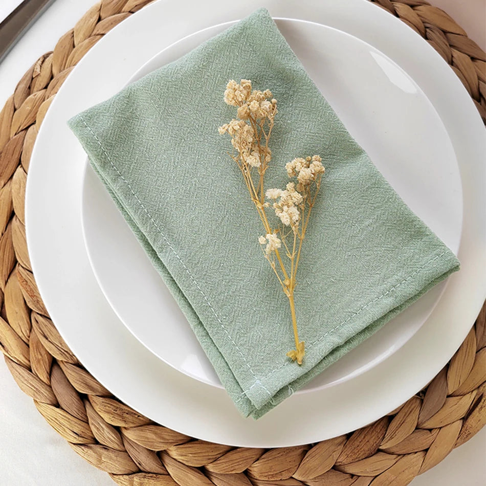 Une serviette verte en coton posée sur une assiette blanche, avec des fleurs sèches disposées dessus, sur un set de table en paille tressée.