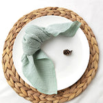 Un ensemble chic et épuré comprenant un napperon en fibres tressées et un chiffon doux de couleur verte pastel, avec un cône de pin brun foncé posé à côté.