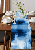 Une table de salle à manger élégamment dressée, avec des assiettes blanches, des verres de vin et un bouquet de fleurs printanières dans un vase, sur un chemin de table bleu et blanc.