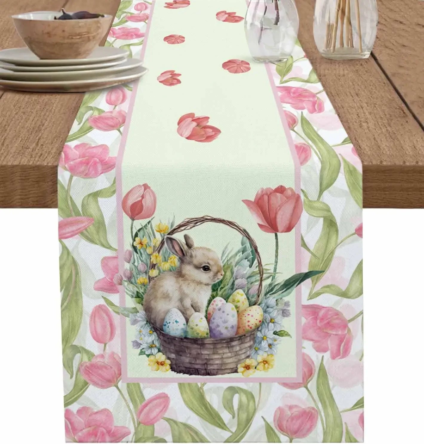 Un petit lapin brun dans un panier avec des œufs de Pâques colorés, entouré de tulipes roses et de feuillage, sur un chemin de table fleuri.