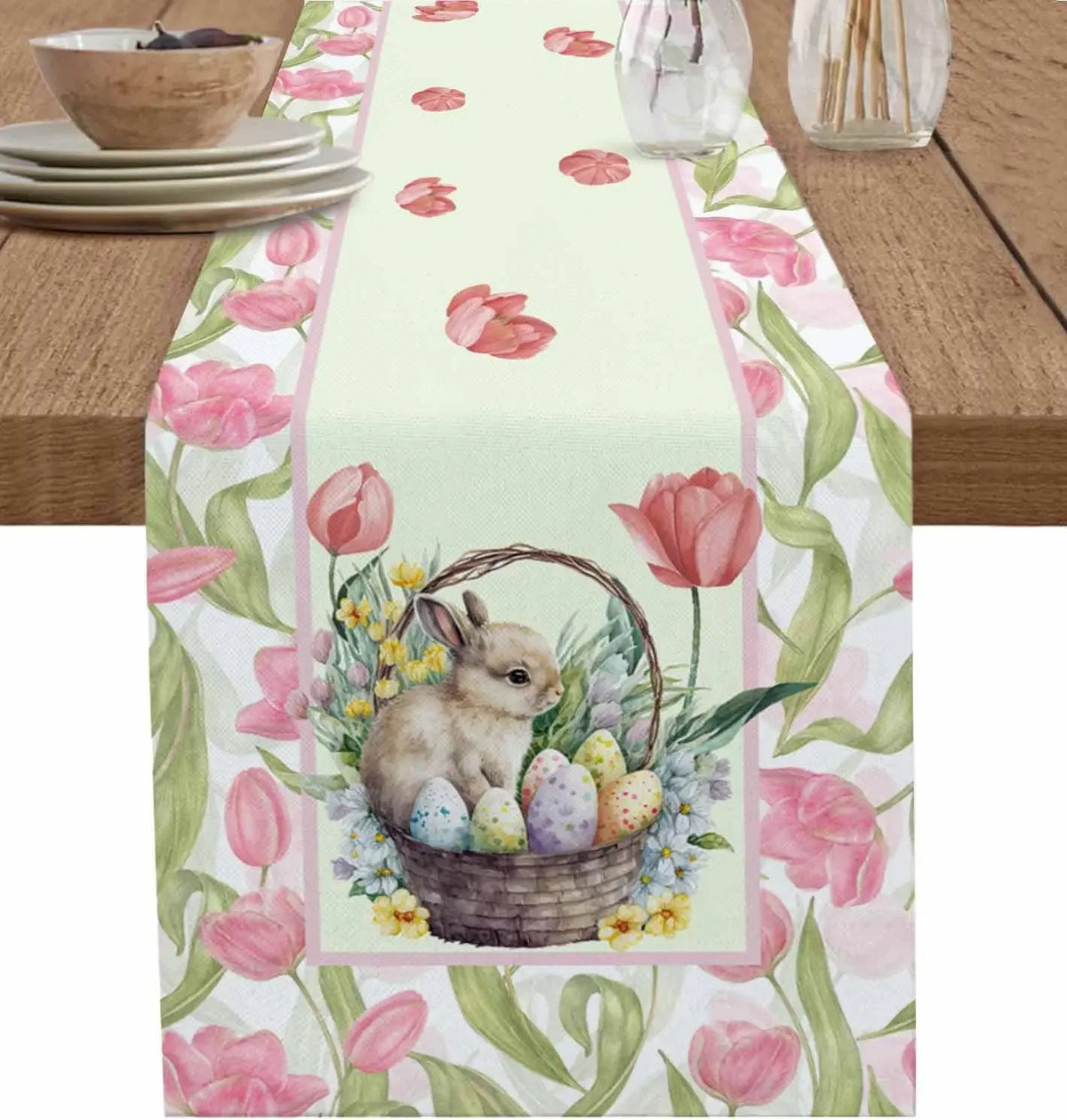 Un panier décoratif en osier contenant un petit lapin et des œufs de Pâques entouré de tulipes roses et de verdure sur un chemin de table blanc.
