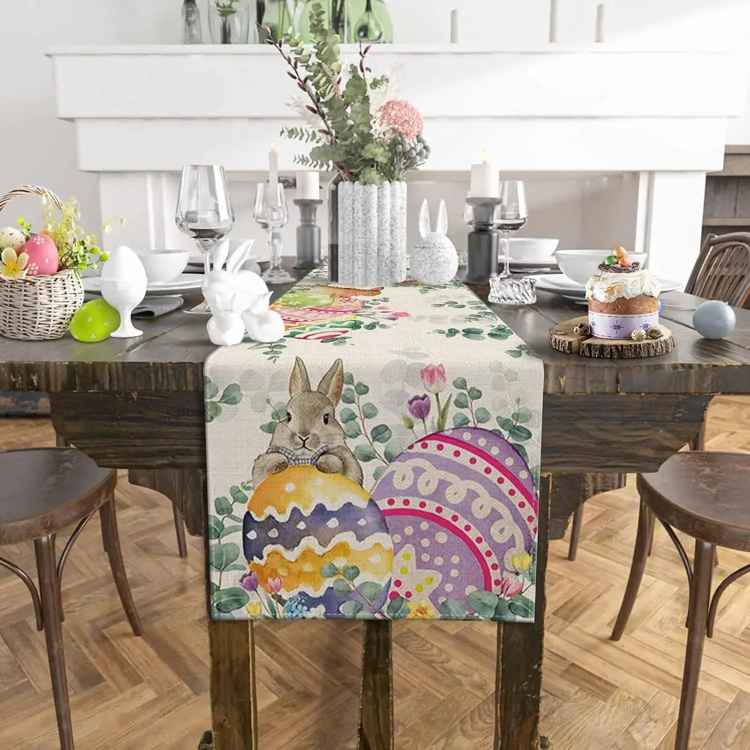 Cette image montre une décoration de table festive avec des éléments de Pâques, notamment des œufs colorés, des lapins en céramique et un centre de table fleuri sur un chemin de table imprimé de motifs de fleurs et de lapins.