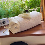 Un sac de couchage cosy et décoré de motifs délicats, posé sur une table en bois avec une horloge vintage en arrière-plan, le tout entouré d'une végétation verdoyante et luxuriante.