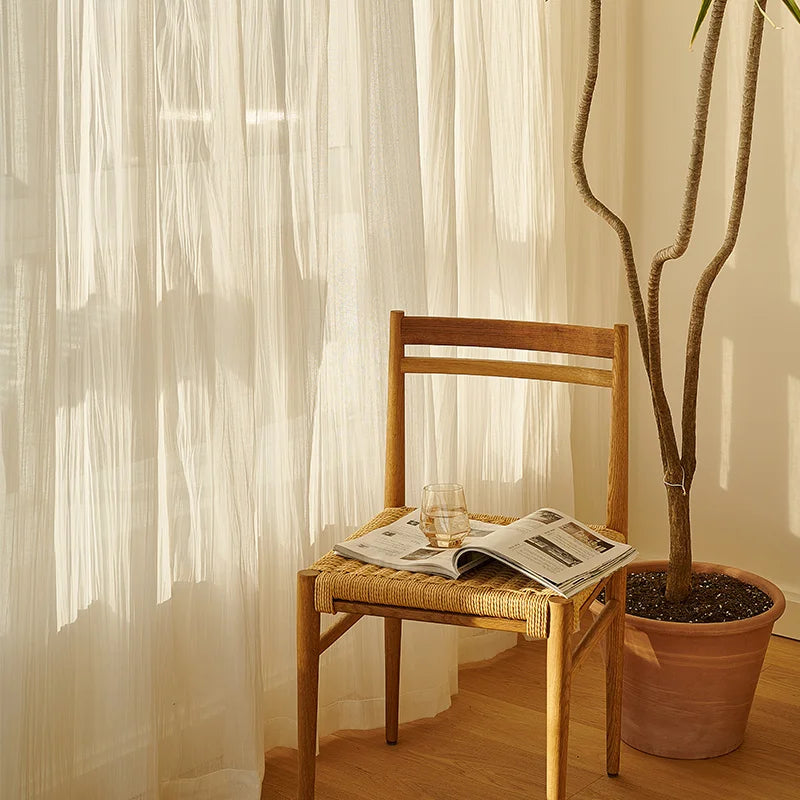 Voilage d'été blanc transparent laissant passer une lumière douce, avec une chaise en bois et un verre posé sur un journal.