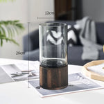 Vase en verre décoratif avec support en bois, mesurant 26 cm de hauteur et 12 cm de diamètre, placé sur une pile de magazines.