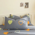 Coussin décoratif gris avec motifs de cœurs orange et verts disposé sur un lit avec des éléments décoratifs muraux.