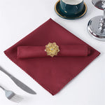 Un mouchoir de table rouge bordeaux avec une fleur décorative en or sur un fond de table blanche avec des ustensiles de table.
