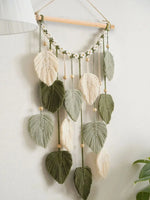 Élégante suspension murale macramé ornée de feuilles vertes et de plumes beiges, créant une décoration naturelle et apaisante.
