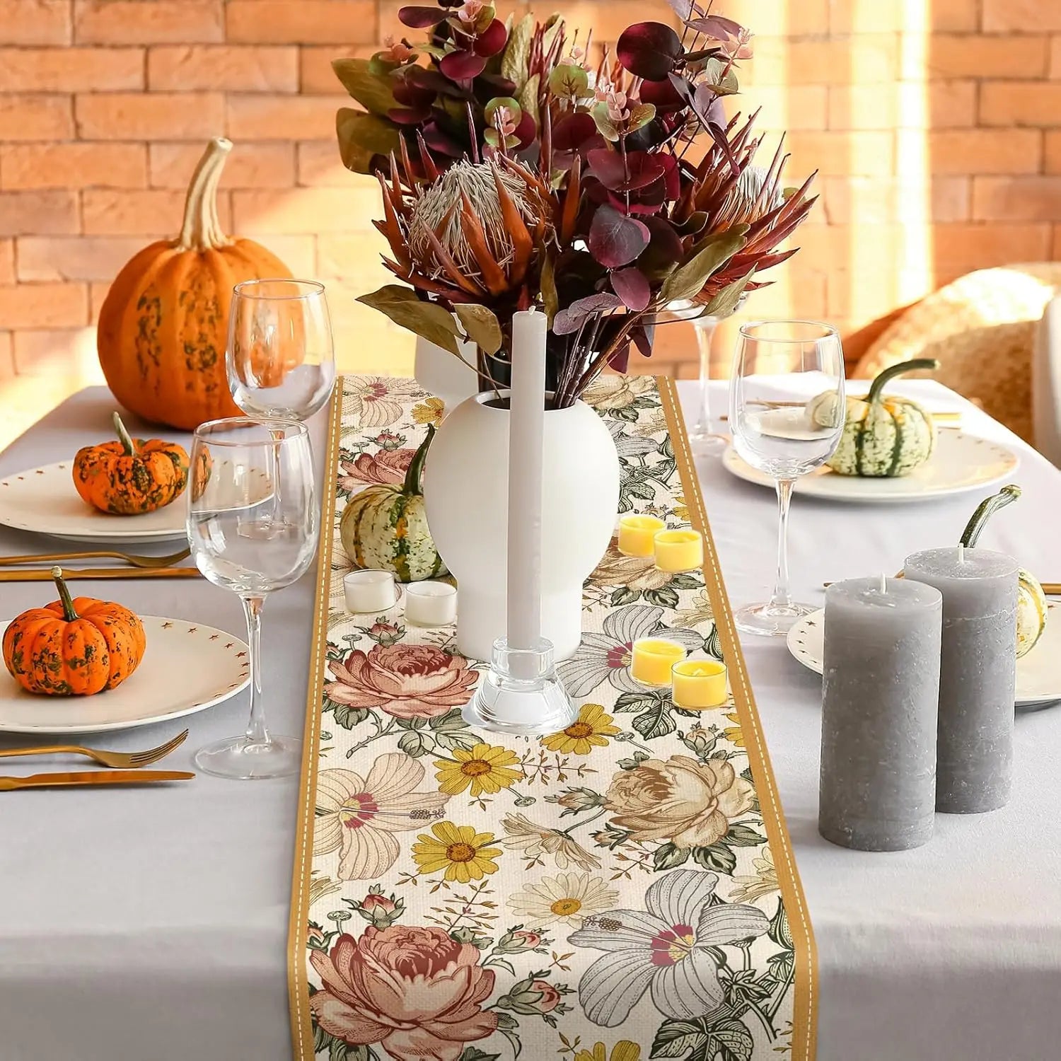 Le centre de table automnal présente un bouquet floral coloré dans un vase blanc, entouré de citrouilles et de verres à vin, sur un chemin de table fleuri.