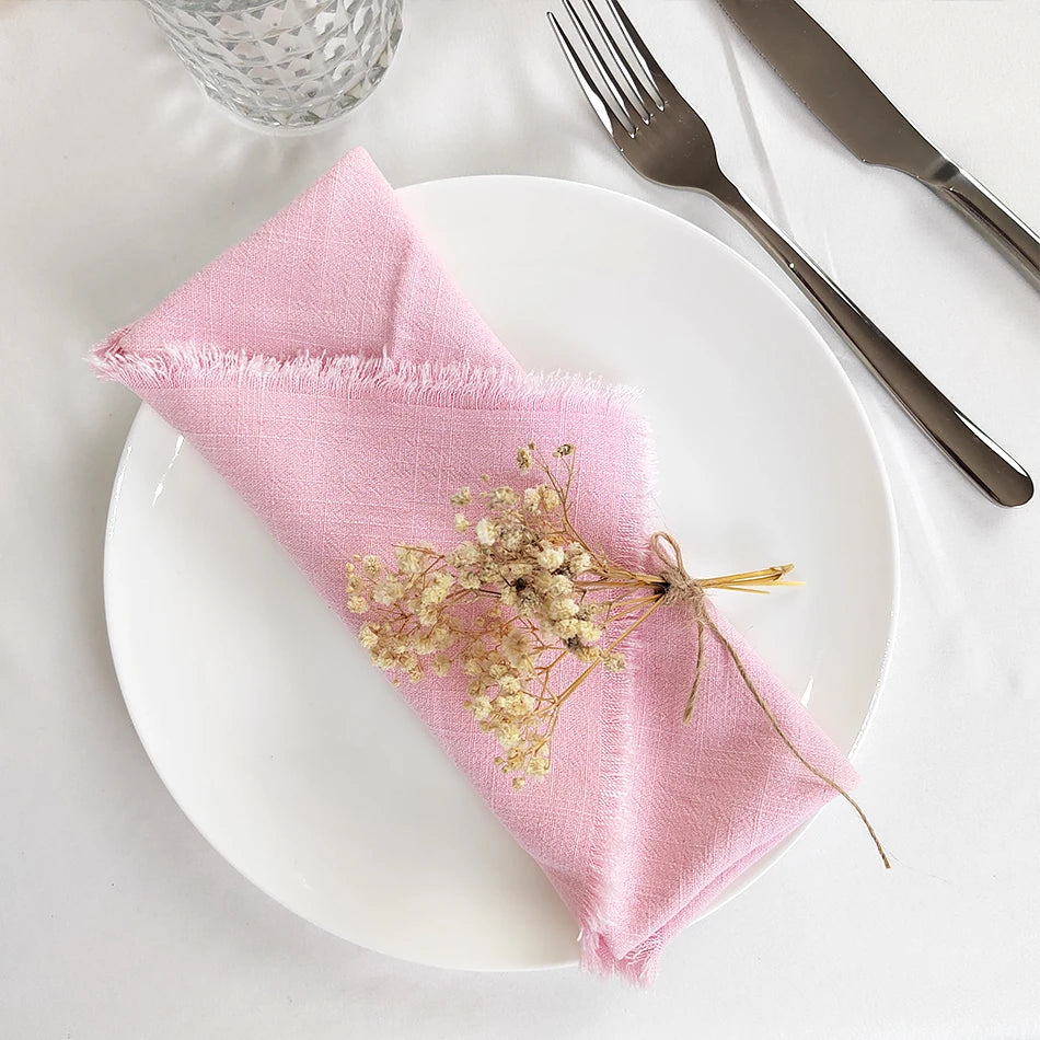Une serviette de table rose pastel, délicatement ornée d'une décoration florale, posée sur une assiette blanche, avec des couverts en métal et un verre en cristal, créant une ambiance élégante et raffinée.