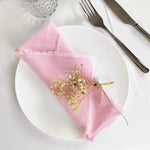 Une serviette de table rose pastel, délicatement ornée d'une décoration florale, posée sur une assiette blanche, avec des couverts en métal et un verre en cristal, créant une ambiance élégante et raffinée.