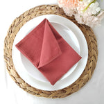 Une serviette de table rouge en tissu posée sur une assiette blanche entourée d'un plateau tressé et de fleurs roses.